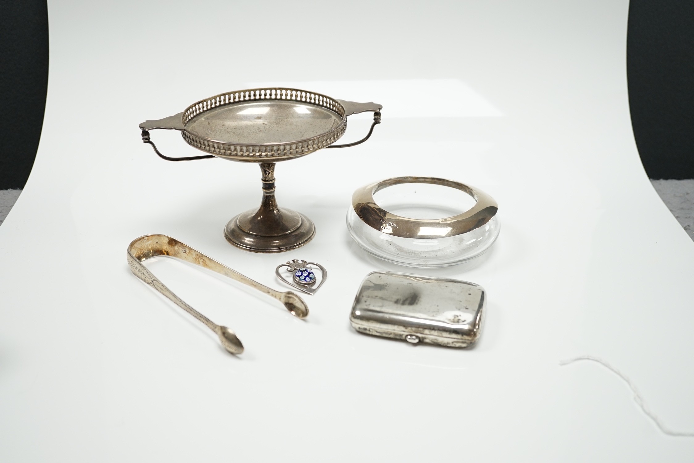 A George V silver tazza, a silver cigarette case, a pair of George III silver sugar tongs, a silver mounted glass shallow dish and a modern silver and lass inset pendant.